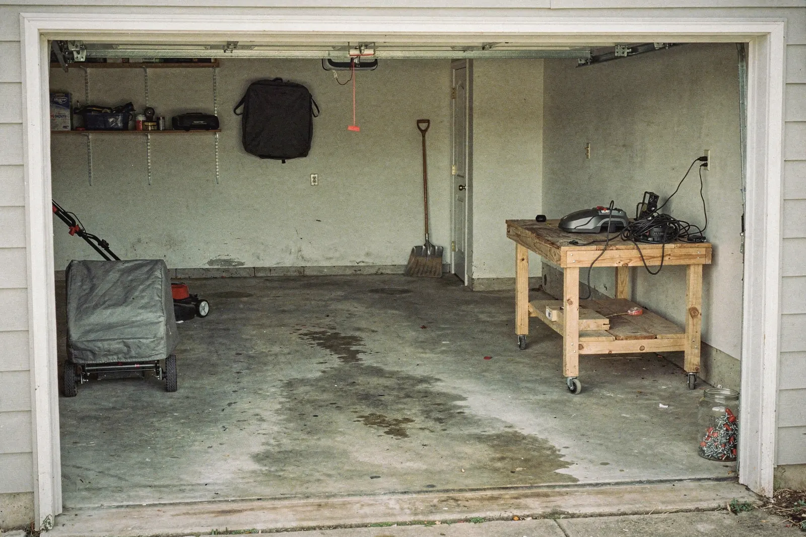 Tidy two-car garage lit from the open door, a mower under a canvas tarp, a workbench with a small charger and a coiled cord, generated illustration