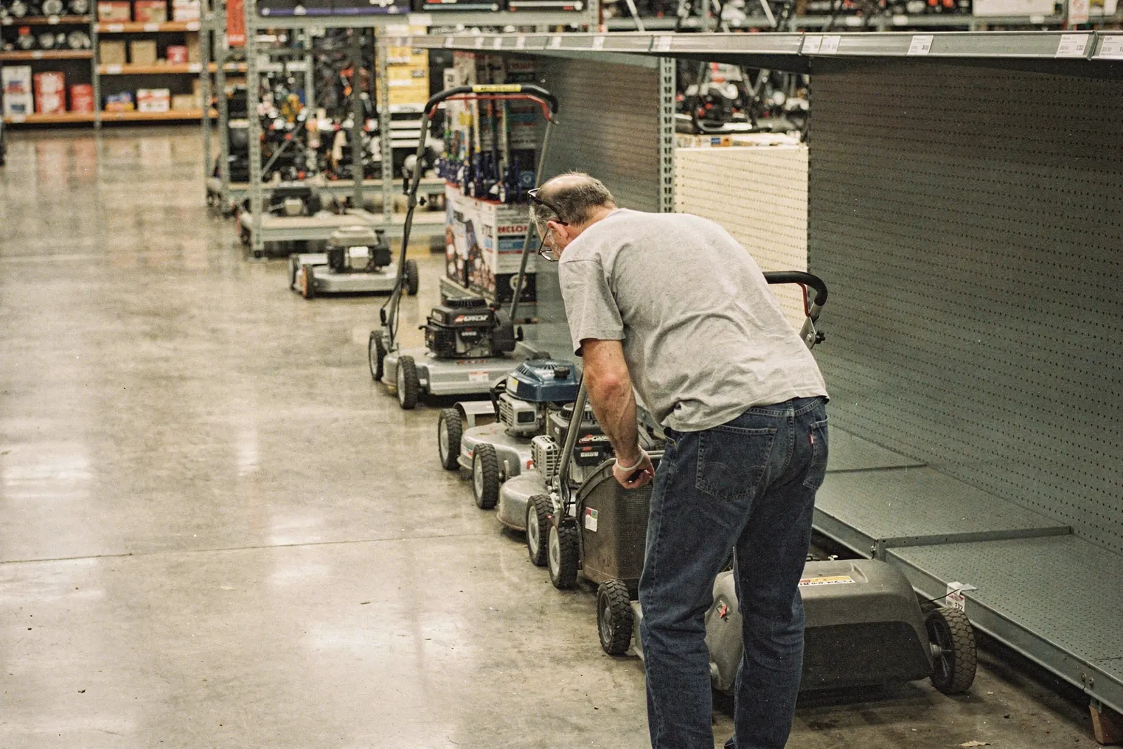 Hardware store lawn aisle with a shopper seen from behind bending to read a blank hang tag on a plain grey walk-behind mower, generated illustration