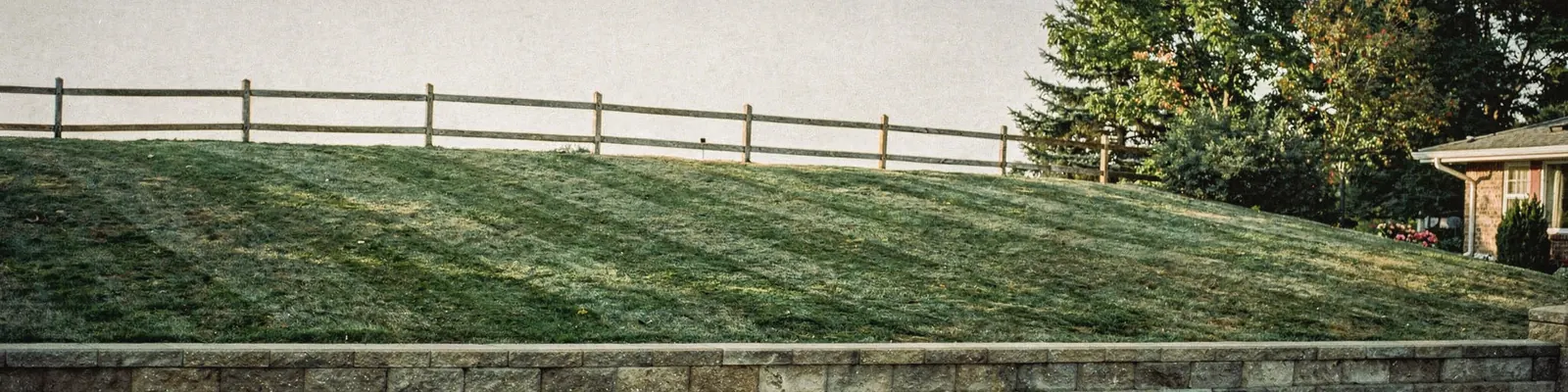 Steep mown grass bank beside a suburban driveway rising from a low stone wall to a split-rail fence in raking evening light, generated illustration
