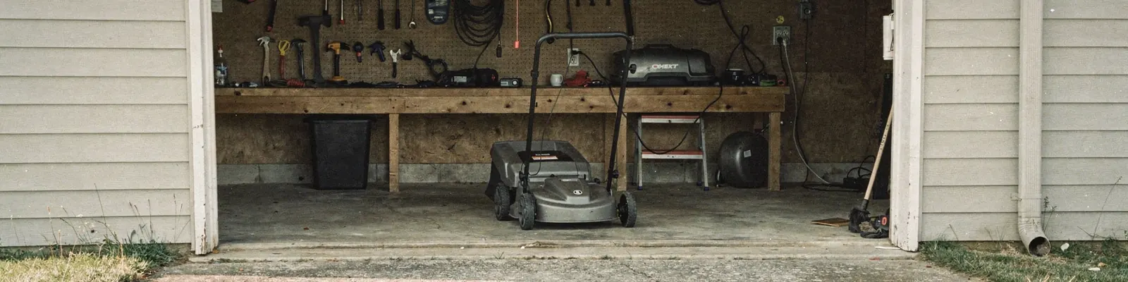 Open suburban garage doorway with a plain charcoal walk-behind mower parked inside, a black battery pack and a small charger on a workbench, generated illustration