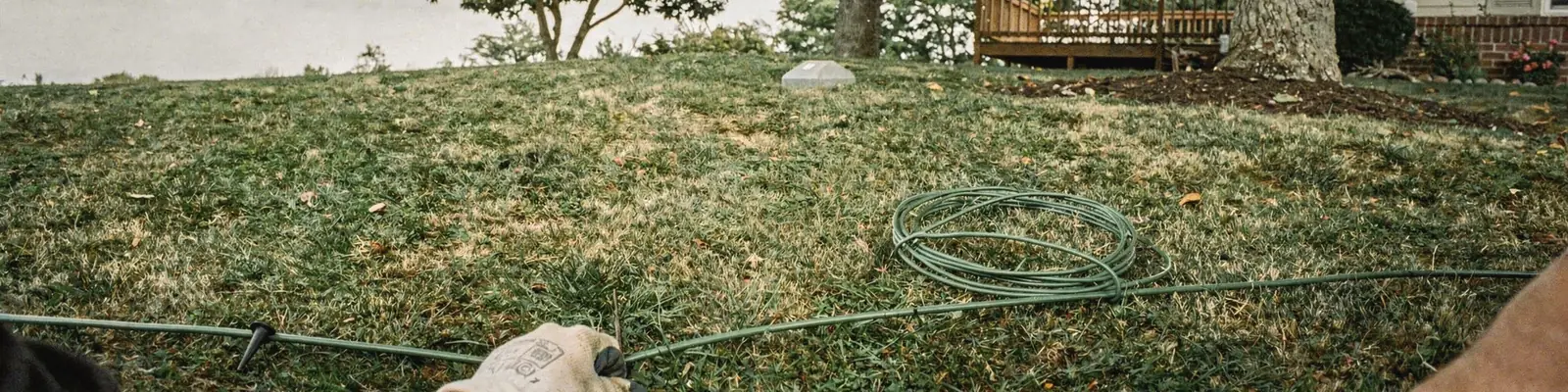 Gloved hands pressing a thin boundary wire into shaded turf with a plastic peg under two oaks, generated illustration